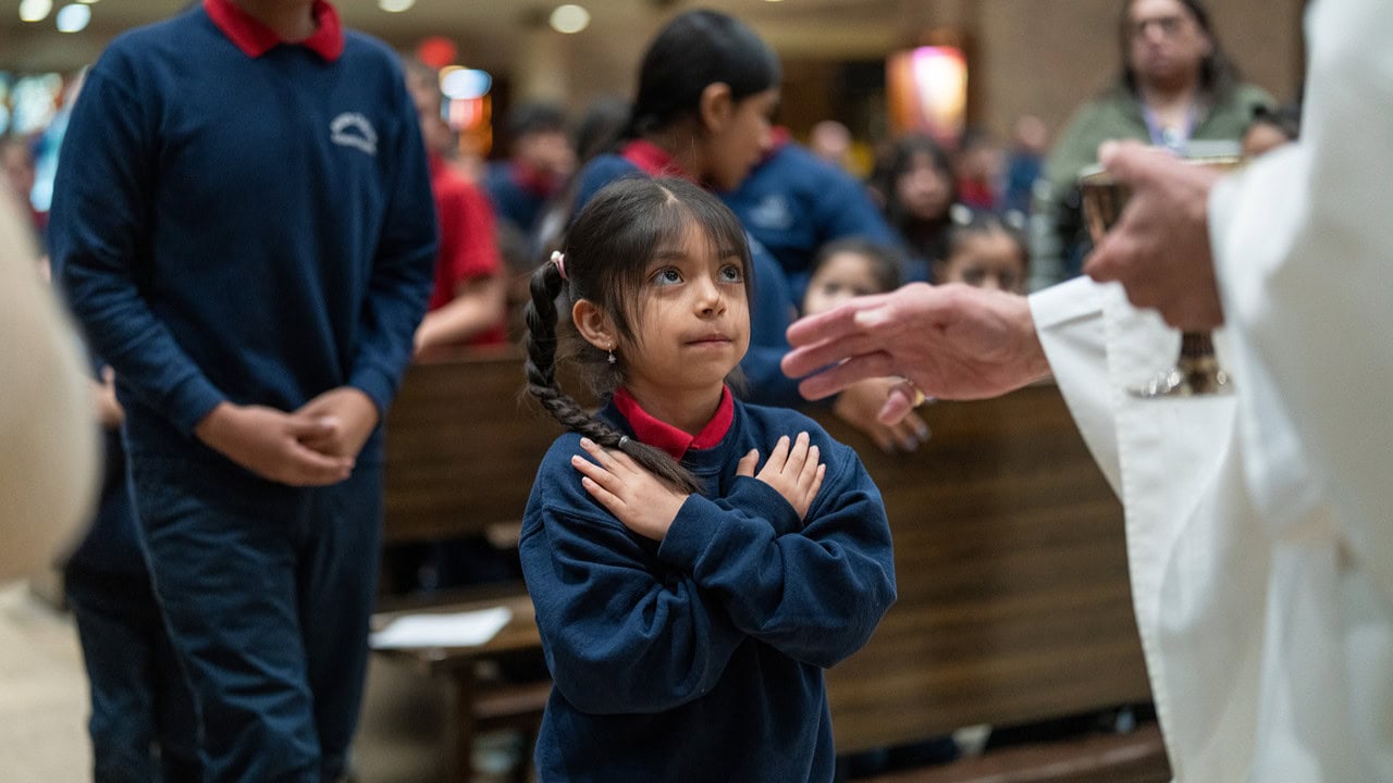 A young Risen Christ student with arms crossed receives a blessing at Mass